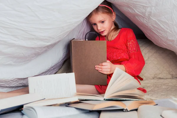 可爱的6 7岁的女孩读一本书 家里的小女孩穿着红衣服躺在床上 盖着毛毯 前面放着许多书 用放大镜搜索书籍中的信息 — 图库照片