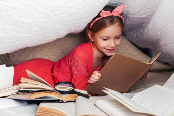 可爱的6 7岁的女孩读一本书 家里的小女孩穿着红衣服躺在床上 盖着毛毯 前面放着许多书 用放大镜搜索书籍中的信息 — 图库照片