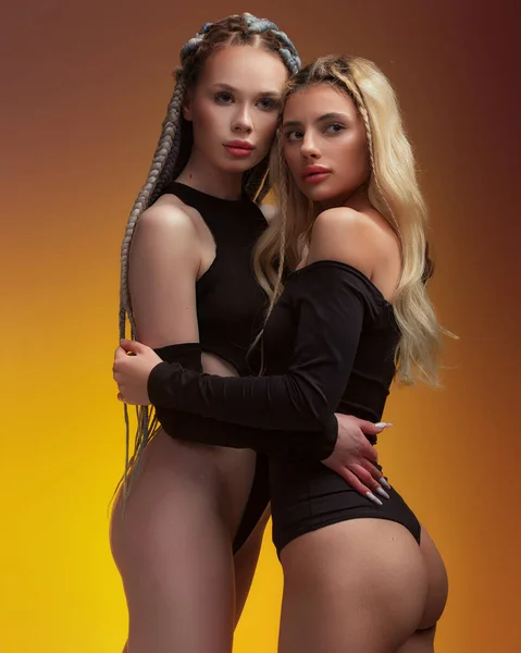stock image Two girlfriend hugging each other, feeling love, posing in black bodysuit with long hair, perfect lips and skin, looking in camera on warm orange background.