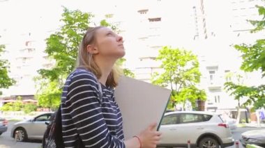 Video alışverişi ya da teslimat konsepti. Güneş gözlüğü takan ve elinde boş bir karton kutu tutan bir kadın şehirde yürüyor.