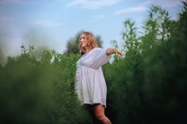 Beyaz boho yaz elbisesi içinde hamile bir kadın uzun yeşil çimlerin üzerinde, tarlanın ortasında yürüyor.