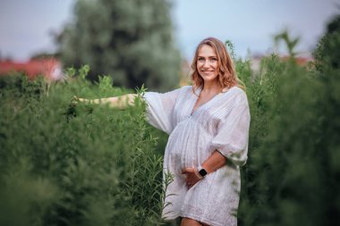 Beyaz boho yaz elbisesi içinde hamile bir kadın uzun yeşil çimlerin üzerinde, tarlanın ortasında yürüyor.