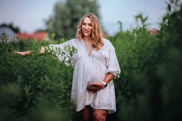 白いBoho夏のドレスで妊娠中の女性は背の高い緑の草の中でフィールドの真ん中を歩く — ストック写真
