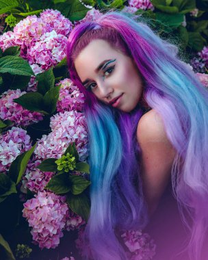 Genç kadın pembe ve menekşe ortanca çiçekleriyle çevrili. Bahçedeki çiçek açan ortancaların üzerine uzanan uzun mor saçlı güzel bir manken.