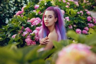 Genç kadın pembe ve menekşe ortanca çiçekleriyle çevrili. Uzun menekşe saçlı güzel bir manken bahçedeki çiçek açan ortancaların tadını çıkarıyor.