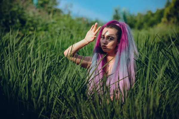 一个有着长长的紫色长发的年轻女人 坐在绿草丛生的草地上 把手举到脸上 挡住了阳光 在他的脸上投下了阴影 — 图库照片