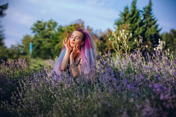 Genç bir kadın, güneşli bir günde lavanta tarlasında oturup rahatça temiz hava alıyor. Uzun menekşe saçlı model kadın.
