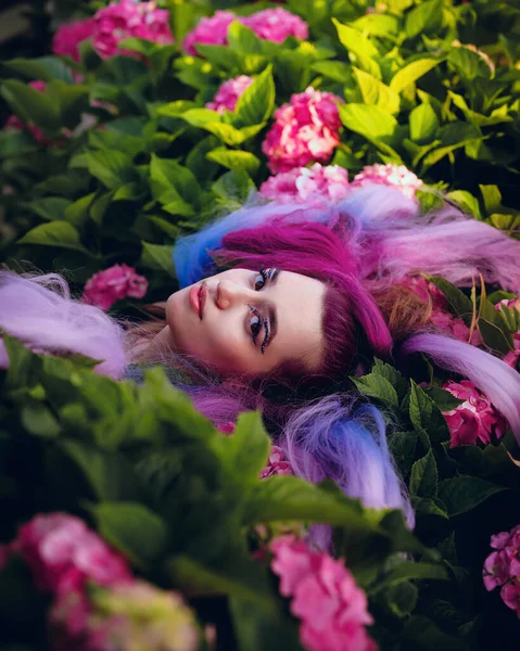 年轻的女人被粉色和紫色的绣花环绕着 美丽的模型 有着长长的彩色紫色头发 花朵盛开在花园里 — 图库照片