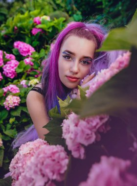 Genç kadın pembe ve menekşe ortanca çiçekleriyle çevrili. Uzun menekşe saçlı güzel bir manken bahçedeki çiçek açan ortancaların tadını çıkarıyor.