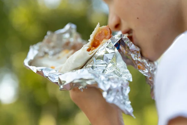 Close up of child mouth eating food in public. Hungry girl bites a big pita or shawarma outdoor.