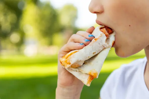Close up of child mouth eating food in public. Hungry girl bites a big pita or shawarma outdoor.