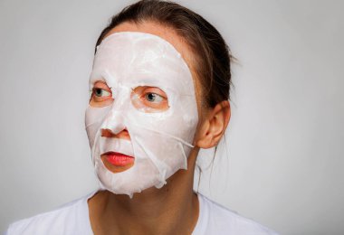 Yüzünde nemlendirici maske olan bir kadın. Kadın yüzüne pamuk yüz nemlendirici maske takıyor, cildine iyi bakıyor, kırışıklıkları önlüyor, izole edilmiş gri arka planda duruyor..