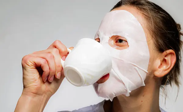 Woman applying cotton facial moisturizing mask on face and drink coffee or tea. Time management and care of skin. Isolated on gray background.
