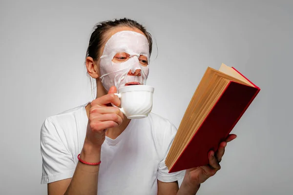女人在脸上涂上棉质面部保湿面膜 喝咖啡 时间管理和皮肤护理 在灰色背景下被隔离 图库图片