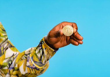 Mavi arka planda duran ve elinde altın bitcoin gösteren renkli gömlekli tanımlanamayan erkek model.