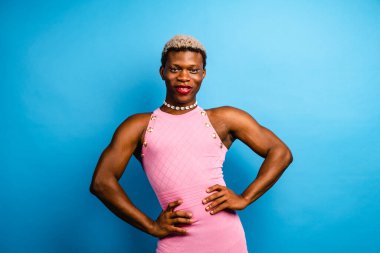 Pembe elbiseli ve kolyeli neşeli, kendinden emin Afrikalı kadın model elleri belinde ve kameraya bakarak mavi duvarın yanında duruyor.