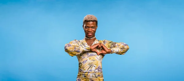 stock image Positive black man in stylish dress looking at camera with smile and showing heart gesture near chest against blue background