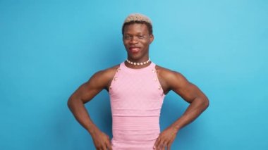 Pembe elbiseli ve kolyeli neşeli, kendinden emin Afrikalı kadın model elleri belinde ve kameraya bakarak mavi duvarın yanında duruyor.