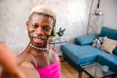 Yukarıdan, pembe elbiseli, saçları boyalı ve dans ederken kameraya bakıp evini gösteren neşeli Afro-Amerikan transseksüel kadından.