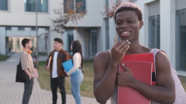 大学のキャンパスに立っているカメラで笑っているアフリカのトランスジェンダーの人のスローモーションビデオ — ストック動画