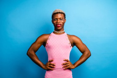 Gülümseyen siyah transseksüel model, boyalı saçlı, pembe elbiseli ve fotoğraf çekimi sırasında elleri belinde mavi arka planda duran aksesuar.