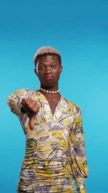 Mide bulandırıcı Afro-Amerikalı şık elbiseli adam başparmağını indiriyor ve mavi arka planda kameraya bakıyor.