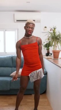 Makyajlı ve kırmızı elbiseli şık bir transseksüel evde sosyal medya için dans ederken kameraya bakıyor. Dikey görüntüler