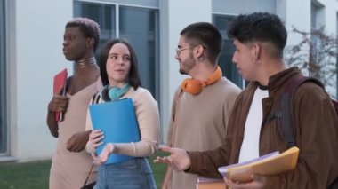 Handheld tracking shot of young woman with folder smiling and speaking with diverse male classmates while walking to studies outside university building together