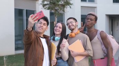 Handheld shot of young Hispanic man in casual clothes using smartphone to take selfie with multiracial classmates grimacing and gesturing V sign after studies outside university building