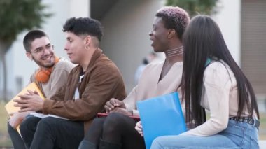 Handheld shot of cheerful black transgender student with dyed hair smiling and covering mouth in amazement while sitting on bench and chatting with diverse classmates after studies