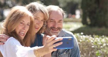 Şehir parkındaki akıllı telefondan video kaydeden neşeli orta yaşlı kadınların ve erkeklerin yavaş çekimleri ekrandaki bulanık arka plana bakarak konuşurken ve sarılırken.