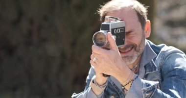 Kot ceketli orta yaşlı bir adamın güneşli bir günde şehir parkındaki eski bir kamerada çekim yapması.