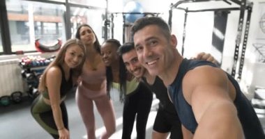 Spor salonunda güneşli bir günde spor yaptıktan sonra neşeli erkek ve kadın sporcularla selfie çekerken aktif kıyafetli mutlu bir yetişkin etnik eğitmen gülümsüyor.