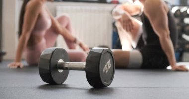 Dumbbell, kimliği belirsiz kaslı erkek sporcunun bulanık geçmişiyle ve spor salonunda spor yaparken yerde oturup sohbet eden aktif giysili formalı bir kadınla yerde bırakılmış.