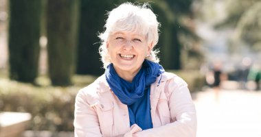 Yaşlı, gri saçlı, kot ceketli, mavi eşarplı bir kadın. El ele tutuşurken kameraya bakıyor ve şehir parkının bulanık arka planına gülümsüyor.