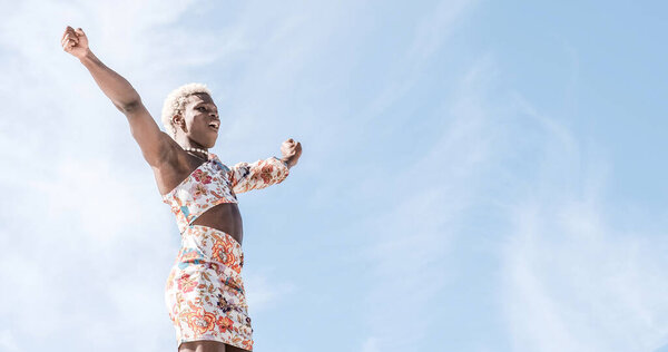 Low angle of carefree young African American transgender person in colorful dress dancing against blue sky and smiling while enjoying freedom on sunny day