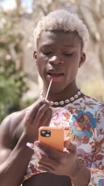 Cinsiyet değiştiren kadın dışarıda dururken cep telefonuna bakarak ruj sürüyor. Makyaj ve güzellik konsepti.