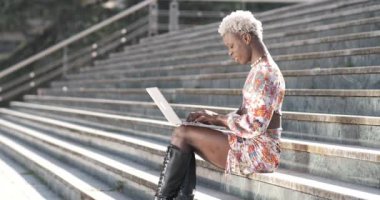 Beton merdivenlerde oturmuş, klavyeyle bilgi yazarken aynı zamanda şehir sokaklarından gelen neşeli, Afro-Amerikan eşcinsel adamın yan görüntüsü.