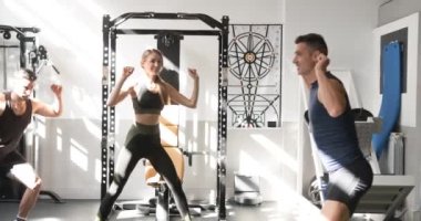 Aktif kıyafetli, gülümseyen kaslı erkek çalıştırıcının yan görünüşü, günışığında modern spor salonunda kararlı, çoklu etnik kökenli bir grup genç sporcuyla yan hamleler yaparken.