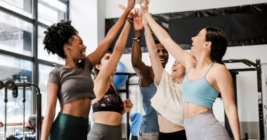Spor kıyafetleri içinde, mutlu bir şekilde gülümseyen ve modern spor salonunda birlikte çalıştıktan sonra beşlik çakan bir grup pozitif, çok kültürlü genç kadın ve erkek.