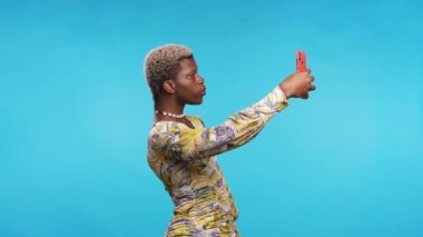 Siyah Afrikalı çift cinsiyetli kişi mavi arka planda selfie çekiyor.
