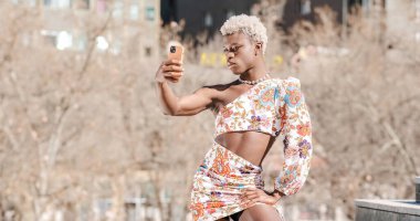 Siyahi elbiseli, Afro-Amerikan bir transseksüel sokakta poz verirken akıllı telefondan kendi portresini çekiyor.