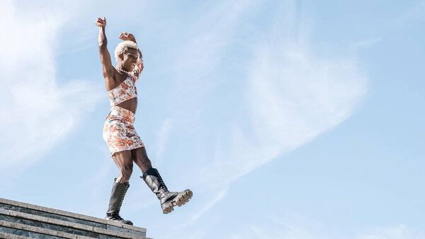 Low angle of carefree young African American transgender person in colorful dress dancing against blue sky and smiling while enjoying freedom on sunny day
