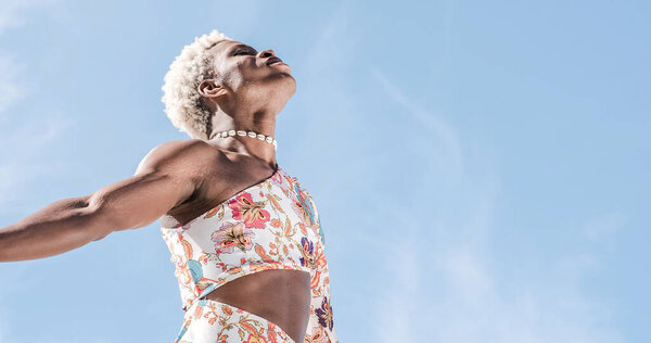 Low angle of carefree young African American transgender person in colorful dress dancing against blue sky and smiling while enjoying freedom on sunny day