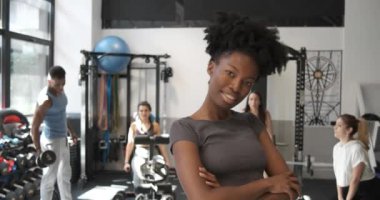 Spor giyimli, kıvırcık saçlı, modern spor salonunda spor yaparken gülümseyen ve kameraya bakan mutlu Afrikalı genç Amerikalı bayan sporcu.
