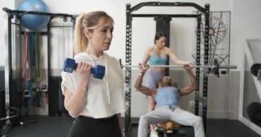 Aktif giyinen sarışın sporcu kadın sporcu yoğun antrenman sırasında ağırlık kaldırarak spor yapıyor. Koça karşı modern spor salonunda halterli zencilere yardım ediyor.