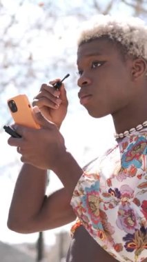 Kirpiklerine maskara sürmüş kıvırcık saçlı, renkli elbiseli ve kolyeli genç Afro-Amerikan transseksüel sokakta akıllı telefon ekranına bakarken