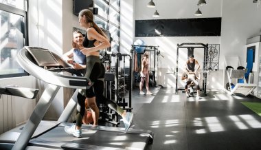 Modern spor salonundaki spor salonunda beden eğitimi dersinde erkek eğitmenle koşu bandında egzersiz yapan neşeli genç bayan sporcu.