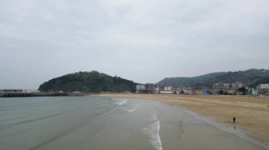 İspanya, Cantabria 'da bulutlu gökyüzüne karşı dalgalı okyanus kenarında yürüyen tanınmayan bir insanın nefes kesici insansız hava aracı görüntüsü