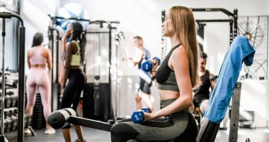 Spor kıyafetli genç bir kadının spor yaparken spor malzemeleriyle ağır halteri kaldırışının yan görüntüsü.